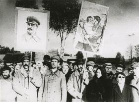 Uczestnicy wiecu z plakatami propagandowymi, na jednym widnieje Józef Stalin, drugi to plakat W. Korieckiego przedstawiający „wyzwolonego” chłopa całującego się z żołnierzem Armii Czerwonej.