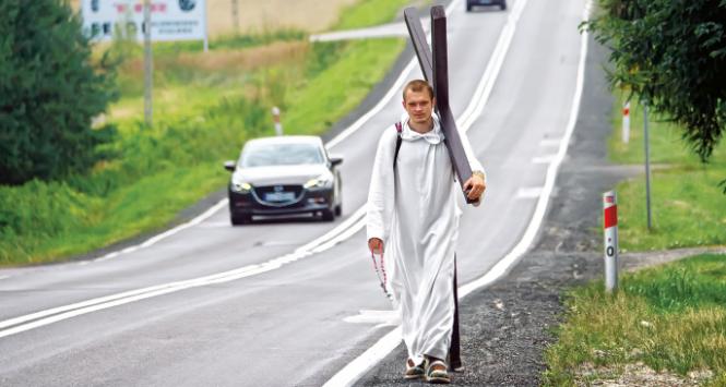 Idzie. Krzyż szura spodem o asfalt, drewno powoli się ściera.