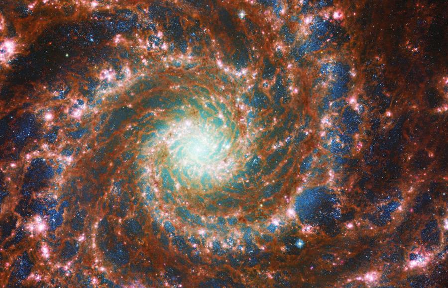 Na tym obrazie, który łączy obserwacje z JWST i Hubble’a, widać efektowne wiry galaktyki M74 (Fantom). Dane zebrane przez Hubble’a w świetle widzialnym pokazują obecne w galaktyce spiralnej gwiazdy. Starsze, bardziej czerwone gwiazdy znajdują się w świecącym jądrze, zaś młodsze, bardziej niebieskie – na obrzeżach galaktyki. Promieniowanie podczerwone uchwycone przez JWST uwypukla z kolei gaz i pył rozmieszczony w ramionach spiralnych, a także jasną gromadę gwiazd w sercu galaktyki. Każdy z teleskopów eksponuje inny aspekt tego kosmicznego cudu, a połączone dane oferują obraz pełniejszy, niż mieliśmy wcześniej.