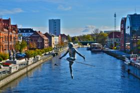 Bydgoszcz ma poza wszystkim swoje symbole. Do najbardziej charakterystycznych należy bezsprzecznie rzeźba Przechodzący przez rzekę. Zawisła 20 metrów nad Brdą na cienkiej linie łączącej dwa brzegi. Jest tutaj od 1 maja 2004 r. Nieprzypadkowo – powstała dla uczczenia rocznicy przystąpienia Polski do Unii Europejskiej. A wydaje się, że jest tu od zawsze, tak bardzo kojarzy się z Bydgoszczą. Praca Jerzego Kędziory waży zaledwie 50 kg, niewiele jak na rzeźbę. Jej środek ciężkości został przeniesiony pod poziom liny, żeby mogła swobodnie balansować, bez obaw, czy na pewno się utrzyma. Figurze towarzyszy jaskółka, z niemieckiego schwalbe – od nazwiska założyciela Filharmonii Pomorskiej Andrzeja Szwalbego.