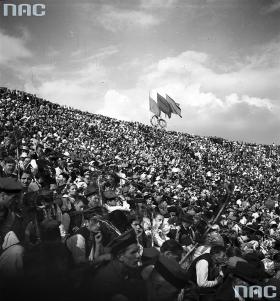 Mozolne wspinanie uwieńczone sukcesem, wszyscy mają miejsca. Czerwień i zieleń flag to nie barwy klubowe ale partyjne. To nie radośnie przebrani kibice na pierwszym planie, lecz zespół regionalny.